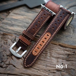 May include: Brown leather watch strap with a perforated design and white stitching. The strap has a silver buckle and a light brown accent piece. The text "No:1" is visible. Designed for an Apple Watch, the strap is shown on a wooden surface.