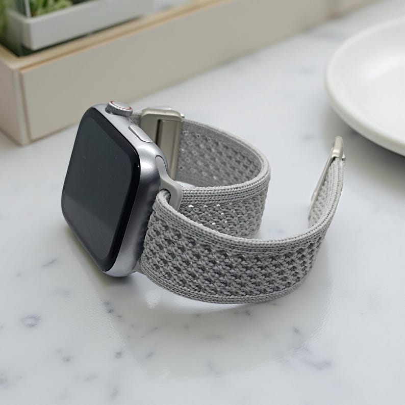 May include: A silver smartwatch with a gray woven band. The watch face is black and the band is a light gray color with a textured pattern. The watch is resting on a white surface.