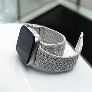 May include: A silver Apple Watch with a gray sport loop band. The band is made of a breathable, woven material with a textured pattern.