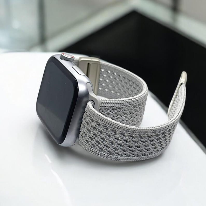 May include: A silver Apple Watch with a gray sport loop band. The band is made of a breathable, woven material with a textured pattern.