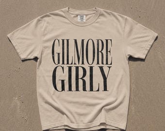 May include: A short-sleeved, crew-neck t-shirt in a light beige color. The front of the shirt features the text GILMORE GIRLY printed in a large, bold, black serif font with a slightly distressed, textured appearance across the center chest.