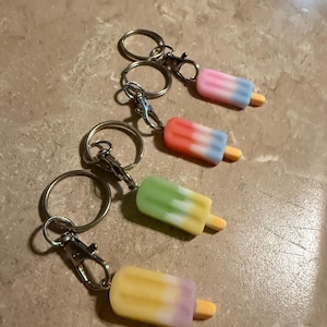 May include: Four colorful popsicle keychains with silver key rings. The popsicles are pink and blue, orange and blue, green and yellow, and yellow and purple.