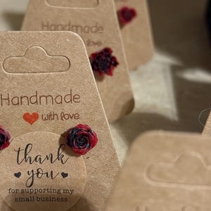 May include: Handmade earrings with a red and black flower design. The earrings are packaged in a brown paper box with a thank you message for supporting small businesses.