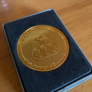 May include: A gold-colored commemorative coin from the Toronto Olympiad, displayed in a black box. The coin features the Olympic rings and text around the edge, including "Centennial Park, Borough of Etobicoke, Ontario, Canada."