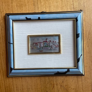 May include: A framed miniature artwork depicting a European town with a bridge, buildings, and trees. The artwork is set within a smaller gold-toned frame, centered on a textured white background. The outer frame is blue with black accents.