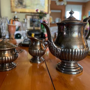 Puede incluir: Un juego de té de plata con tetera, jarra de crema y azucarero. El juego tiene un diseño clásico con un patrón acanalado y asas ornamentadas.