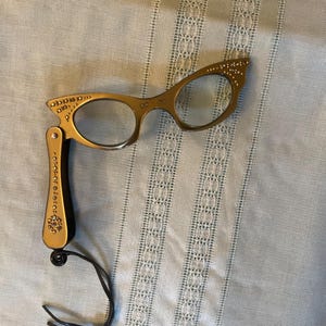Op de afbeelding: Goudkleurige leesbril in vintage-stijl met een cat-eye vorm. De bril heeft decoratieve versieringen en een opvouwbaar handvat. Een zwart koord is aan het handvat bevestigd. Op een lichtblauwe achtergrond.