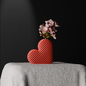Könnte beinhalten: Eine herzförmige, korallenfarbene Vase mit diagonalen Rillen, die einen Strauß rosa Blumen enthält. Die Vase steht auf einer weißen Oberfläche vor einem dunklen Hintergrund und erzeugt einen auffälligen Kontrast.