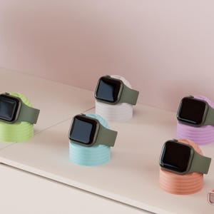 Könnte beinhalten: Sechs Smartwatches mit olivgrünen Armbändern werden auf farbenfrohen, zylindrischen Ständern präsentiert. Die Ständer sind in Pastelltönen von Grün, Blau, Weiß, Lila und Pfirsich gehalten. Die Uhren haben schwarze Bildschirme.