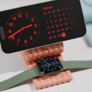 Könnte beinhalten: Ein schwarzes Smartphone zeigt eine rote Uhr und einen Augustkalender, die auf einem rosa Ständer liegen. Darunter wird eine grüne Smartwatch mit dunklem Zifferblatt angezeigt. Tech-Zubehör.