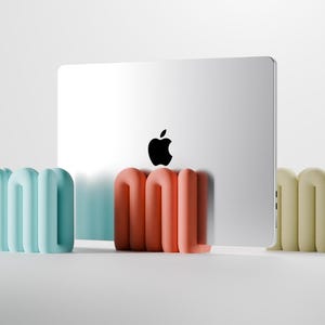 Könnte beinhalten: Ein silberner Laptop mit schwarzem Apple-Logo steht hinter drei farbenfrohen, abstrakten, gebogenen Ständern. Die Ständer sind in Hellblau, Koralle und Hellgelb gehalten. Die Objekte stehen auf einer weißen Oberfläche.
