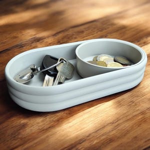 Könnte beinhalten: Eine hellgraue, zweigeteilte Ablage mit abgerundeten Kanten. Ein Fach enthält Schlüssel, das andere Münzen. Die Ablage steht auf einer Holzoberfläche. Schlüssel und Münzen sind sichtbar. Schlichtes, modernes Design.