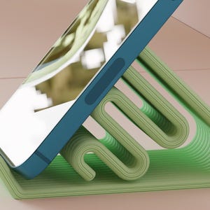 Könnte beinhalten: Ein türkisfarbenes Smartphone liegt auf einem hellgrünen, abstrakten Telefonständer. Der Ständer hat ein geschwungenes, geschichtetes Design mit einer strukturierten Oberfläche. Der Bildschirm des Telefons spiegelt die Umgebung wider. Der Ständer ist für ein Telefon konzipiert.