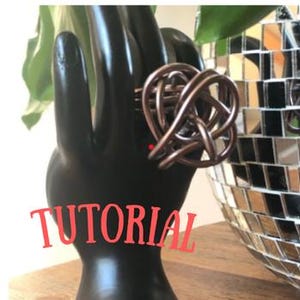 May include: A copper-colored wire ring with a Celtic knot design is displayed on a black hand-shaped jewelry holder. The word "TUTORIAL" is overlaid in red script. A disco ball is in the background.