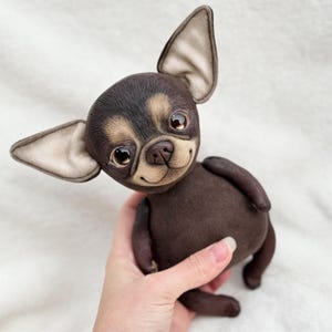 Può includere: Un giocattolo di peluche a forma di chihuahua marrone e beige con grandi occhi marroni e un viso sorridente. Il giocattolo è fatto di tessuto morbido e ha un aspetto realistico.
