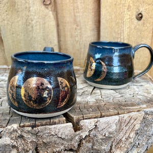 May include: Two ceramic mugs with a dark blue glaze and gold moon phase designs. The mugs have a rounded shape and a handle. They are sitting on a weathered wooden surface.