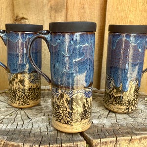 May include: Three ceramic travel mugs with black lids and handles. The mugs have a blue and purple glaze with a mountain and tree design. The mugs are sitting on a wooden surface.