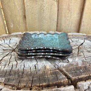 May include: A stack of four small, rectangular ceramic dishes with a teal glaze and a floral pattern. The edges are scalloped and the rims are a dark brown. The dishes are stacked on a weathered wooden surface.