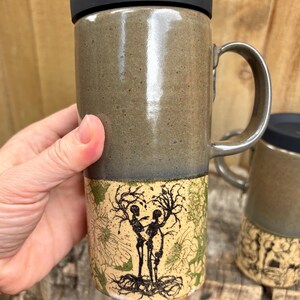 May include: A tall ceramic travel mug with a black silicone lid and a handle. The upper portion is a smooth, speckled olive-green glaze, while the lower section features a cream-colored base with a black ink illustration of two skeletons.