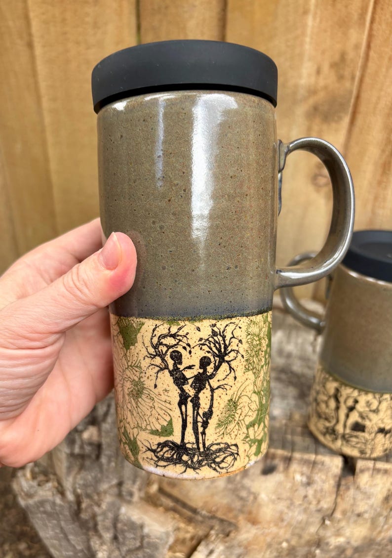 May include: A tall ceramic travel mug with a black silicone lid and a handle. The upper portion is a smooth, speckled olive-green glaze, while the lower section features a cream-colored base with a black ink illustration of two skeletons.