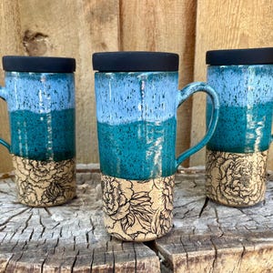 May include: Three tall ceramic mugs with black lids, each featuring a gradient of light blue and teal glaze over a speckled finish, with a base section decorated in a black line-art floral pattern of peonies on a light beige clay background.
