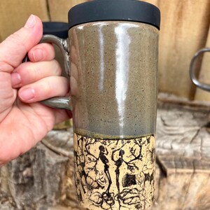 May include: A tall ceramic travel mug with a black silicone lid and a handle. The upper portion features a smooth, speckled olive-green glaze, while the lower section displays a detailed black ink illustration of two skeletons amidst floral vines.