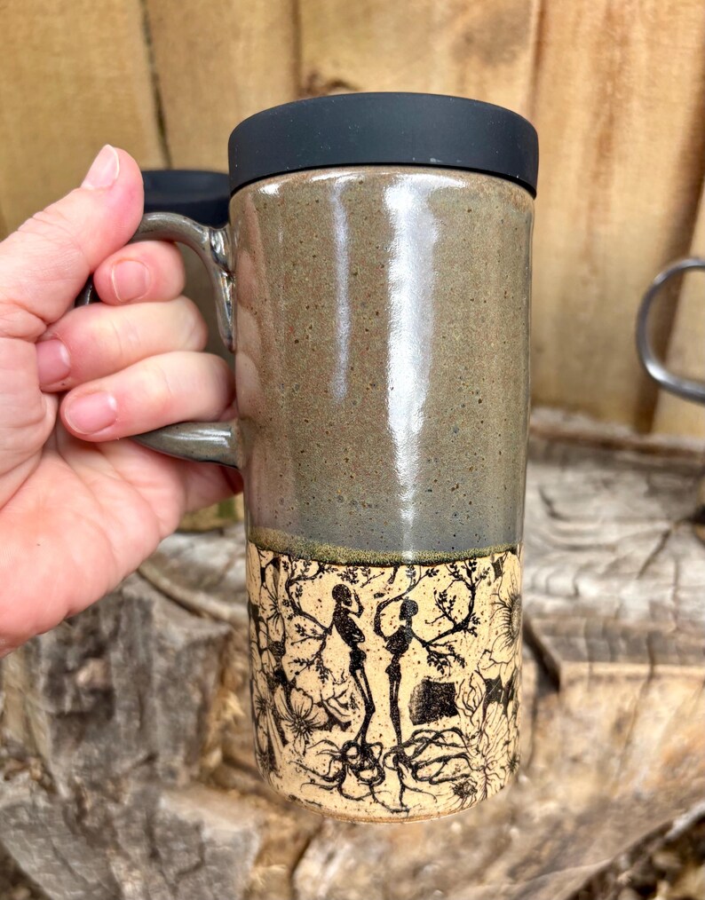 May include: A tall ceramic travel mug with a black silicone lid and a handle. The upper portion features a smooth, speckled olive-green glaze, while the lower section displays a detailed black ink illustration of two skeletons amidst floral vines.