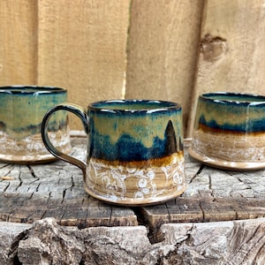 May include: Three handmade ceramic mugs with a unique glaze. The mugs feature a gradient of colors, including blue, green, and brown, with a textured white pattern near the base. Each mug has a brown handle.