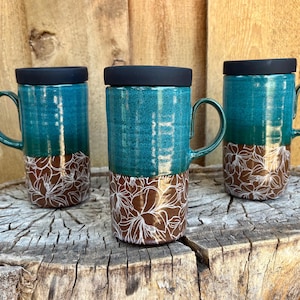 May include: Three tall ceramic mugs with black lids and handles. The upper portion of each mug is a glossy teal blue, while the lower portion is a dark brown featuring a white line-art floral pattern of overlapping petals and leaves.