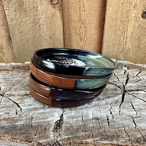 May include: A stack of four handmade ceramic dishes. The top dish is black, the second is brown, the third is green and brown, and the bottom dish is dark brown and blue. The dishes are stacked on a weathered wooden surface.