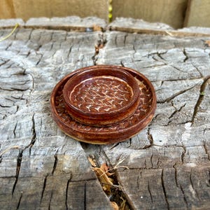 May include: A handmade, brown ceramic bowl and saucer set. The bowl sits atop the saucer, both featuring a textured, leaf-like pattern. The set is displayed on a weathered, wooden surface, showcasing the natural tones of the pottery.