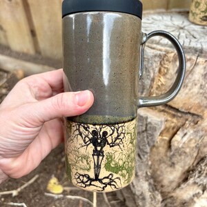 May include: A tall ceramic travel mug with a black lid and a handle. The top half is a smooth, speckled dark gray glaze, while the bottom half features a cream-colored base with a black ink illustration of two skeletons intertwined in a tree.