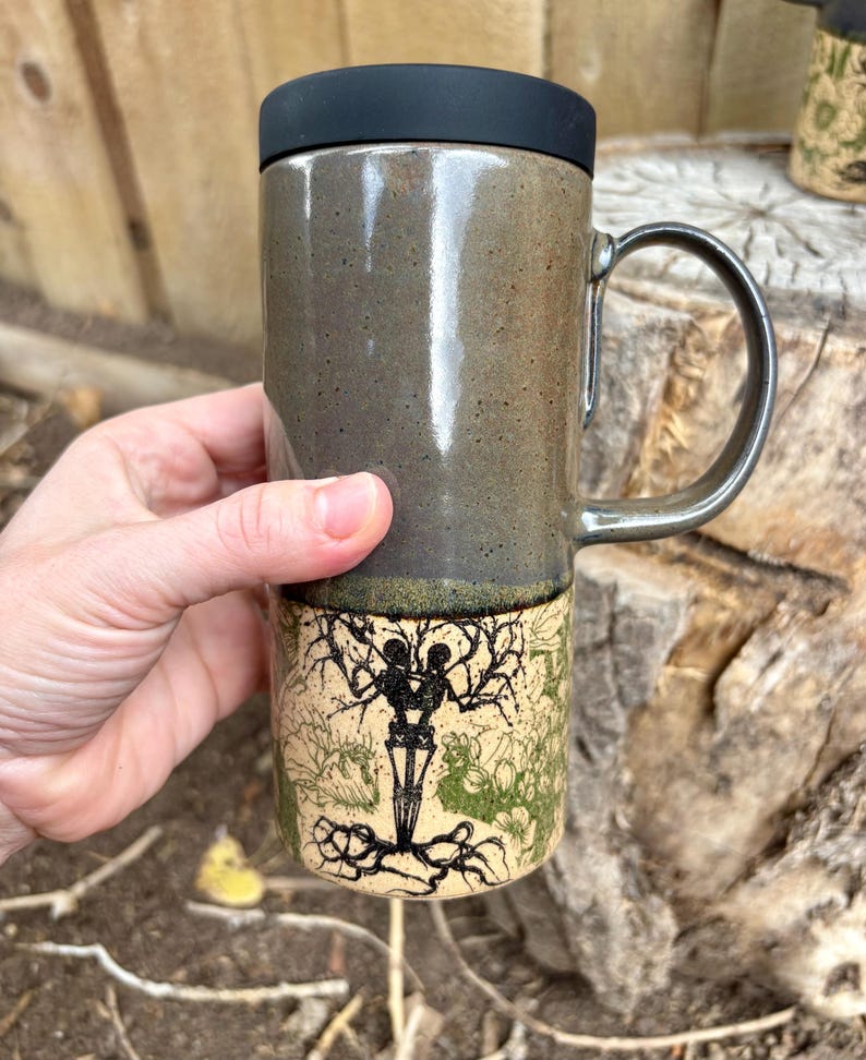 May include: A tall ceramic travel mug with a black lid and a handle. The top half is a smooth, speckled dark gray glaze, while the bottom half features a cream-colored base with a black ink illustration of two skeletons intertwined in a tree.