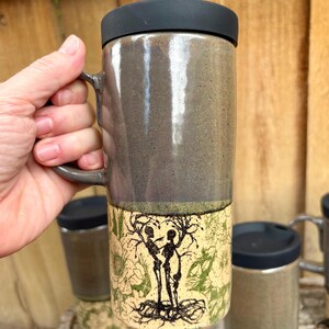 May include: A tall ceramic travel mug with a black lid and a handle. The upper portion has a smooth, glossy dark gray glaze, while the lower portion features a cream-colored background with a black ink illustration of two skeletons and trees.