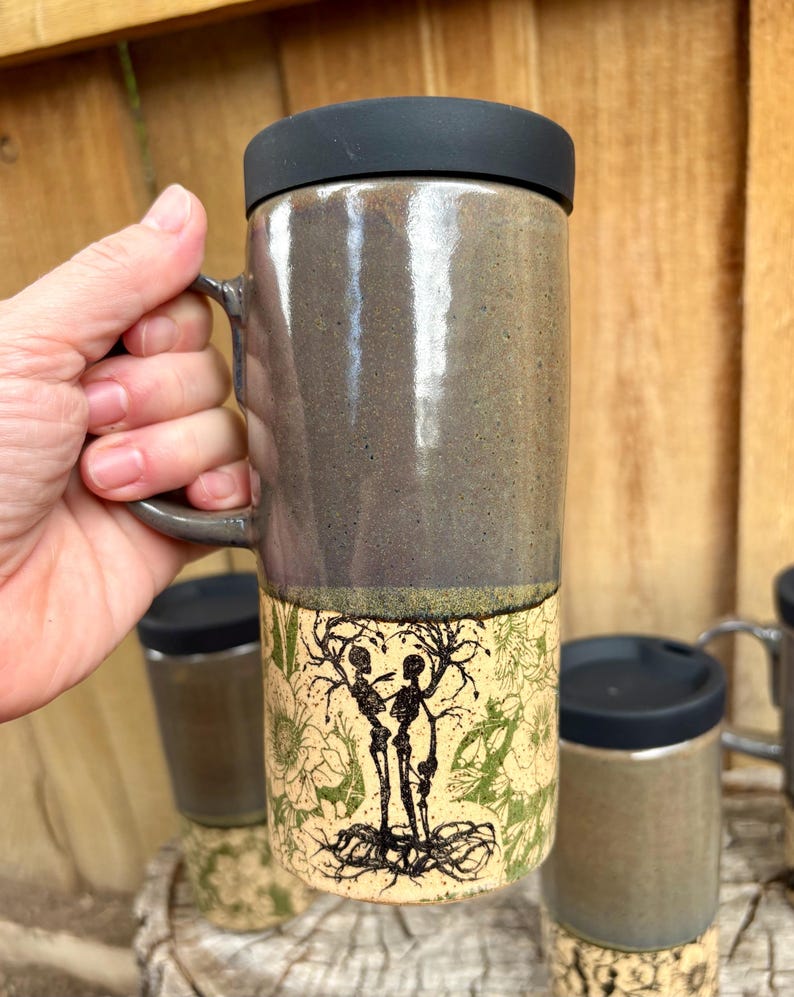 May include: A tall ceramic travel mug with a black lid and a handle. The upper portion has a smooth, glossy dark gray glaze, while the lower portion features a cream-colored background with a black ink illustration of two skeletons and trees.