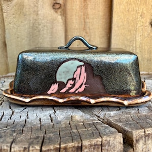 May include: A rectangular ceramic butter dish with a dark, metallic-flecked glaze and a central handle. The front features a hand-painted design of a pale moon rising over a dark, jagged mountain silhouette in shades of pink and brown.