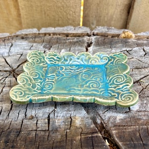 May include: A rectangular ceramic dish with a turquoise interior and a scalloped edge. The dish features a swirling cloud design and is finished in shades of green and blue. The dish is placed on a weathered wooden surface.