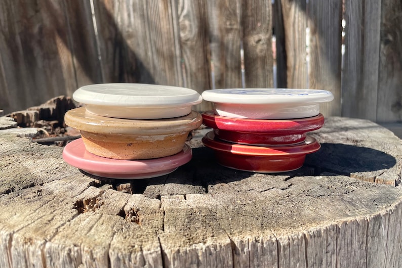 May include: Two stacks of ceramic lids, each featuring a flat circular top and a slightly smaller base. The left stack includes a white, a tan, and a pink lid. The right stack consists of a white, a dark red, and a terracotta-colored lid.