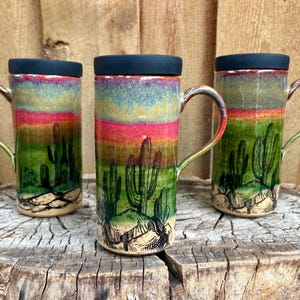 May include: Three tall ceramic mugs with black lids, each featuring a hand-painted desert landscape with green cacti, rocky ground, and a vibrant sunset sky gradient of green, orange, and pink colors, complete with a curved handle on each.