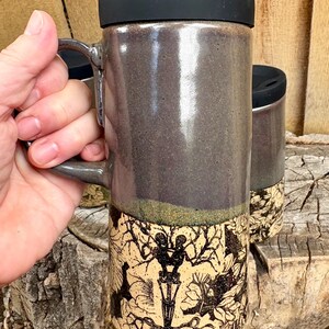 May include: A tall ceramic travel mug with a black lid and a handle. The upper portion is a smooth, dark gray glaze, while the lower section features a cream-colored base with a detailed black ink illustration of two skeletons among flowers.