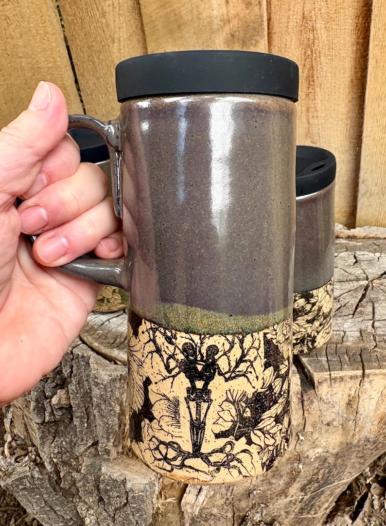 May include: A tall ceramic travel mug with a black lid and a handle. The upper portion is a smooth, dark gray glaze, while the lower section features a cream-colored base with a detailed black ink illustration of two skeletons among flowers.