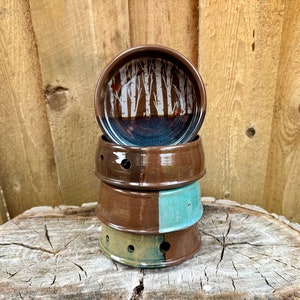May include: Three stacked ceramic bowls with a brown glaze. The top bowl has a white and brown design of birch trees. The middle bowl has a brown glaze with a row of holes around the top edge. The bottom bowl has a brown glaze on one side and a blue glaze on the other side.