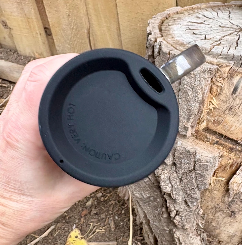 May include: A black circular travel mug lid featuring a small drinking aperture and the embossed text CAUTION: VERY HOT. The lid is constructed from a matte black material and is designed to fit securely onto the top of a beverage container.