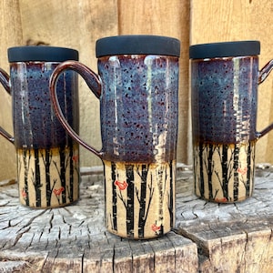 May include: Three tall ceramic travel mugs with black lids and handles. The upper portion features a speckled blue and purple glaze, while the lower section displays a painted forest of black birch trees with small red birds on a cream background.