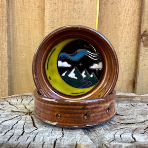 May include: Two-piece ceramic coaster set. The top coaster features a night scene with a yellow crescent moon, white clouds, and green mountains. The coasters are brown with a glossy finish and are stacked on top of each other.