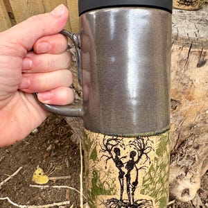 May include: A tall ceramic travel mug with a glossy dark gray upper section and a matte beige lower section. The lower portion features a black ink illustration of two skeletons intertwined with tree branches and roots, accented by green foliage.