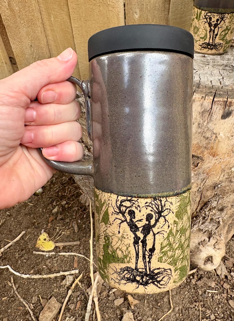 May include: A tall ceramic travel mug with a glossy dark gray upper section and a matte beige lower section. The lower portion features a black ink illustration of two skeletons intertwined with tree branches and roots, accented by green foliage.