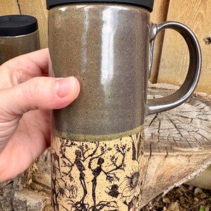 May include: A tall ceramic travel mug with a black silicone lid and a handle. The upper portion has a smooth, glossy dark gray glaze, while the lower portion features a matte cream finish with a detailed black ink illustration of skeletons.