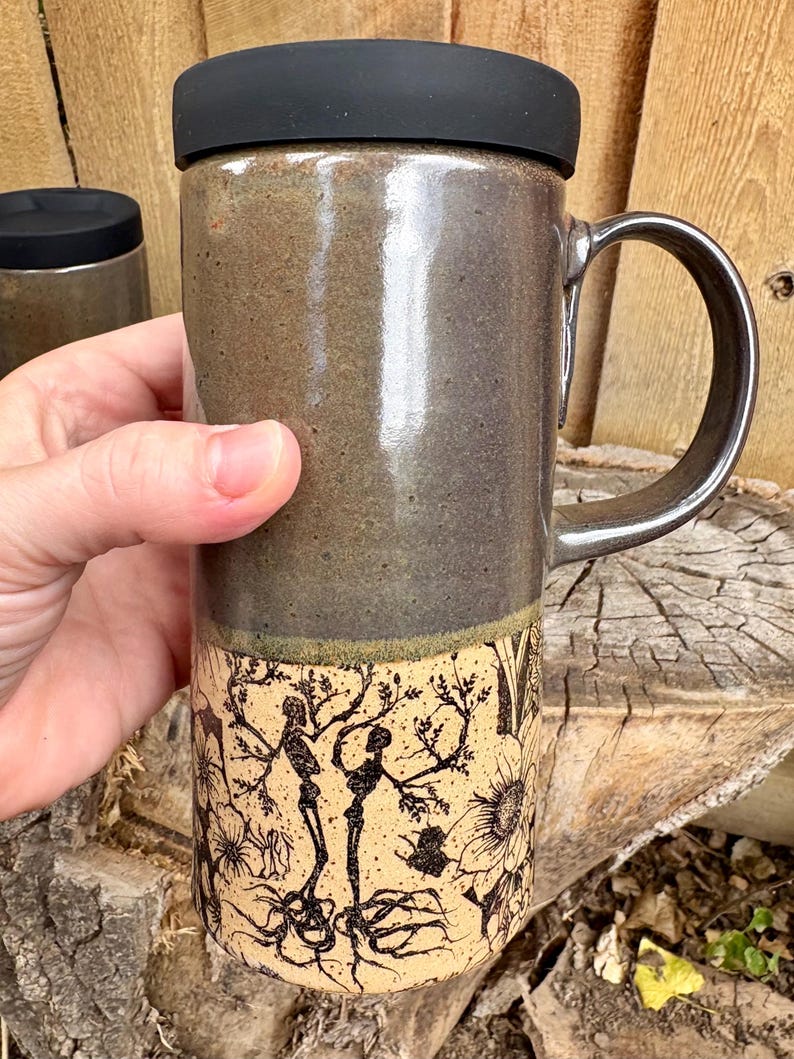 May include: A tall ceramic travel mug with a black silicone lid and a handle. The upper portion has a smooth, glossy dark gray glaze, while the lower portion features a matte cream finish with a detailed black ink illustration of skeletons.