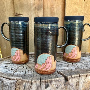 May include: Three tall ceramic mugs with black lids and handles. Each mug features a dark, glossy glaze with a hand-painted desert landscape at the base, showing pink rock formations against a light blue sky, set on a textured wooden surface.